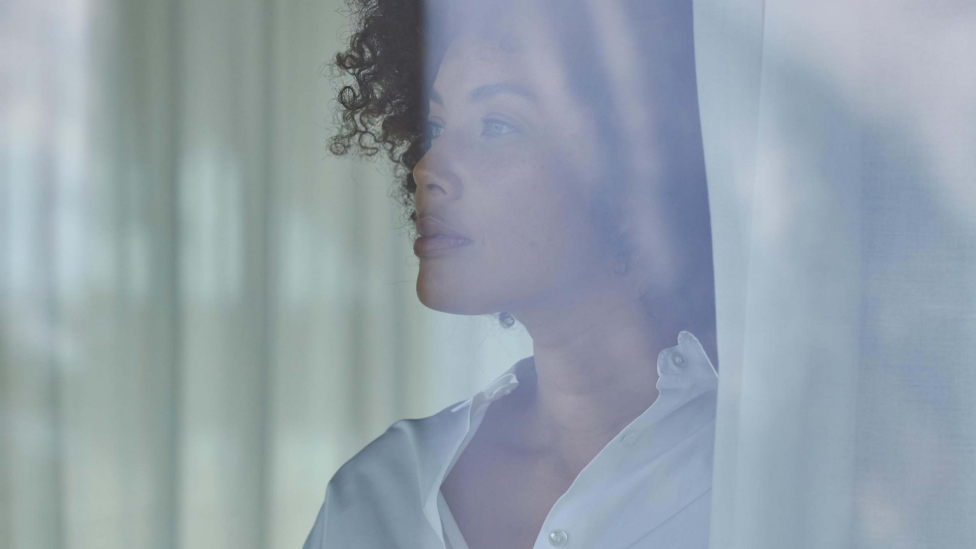 Your hotel in Val Pusteria/Pustertal: Olangerhof Woman in white shirt looking out the window through a curtain.