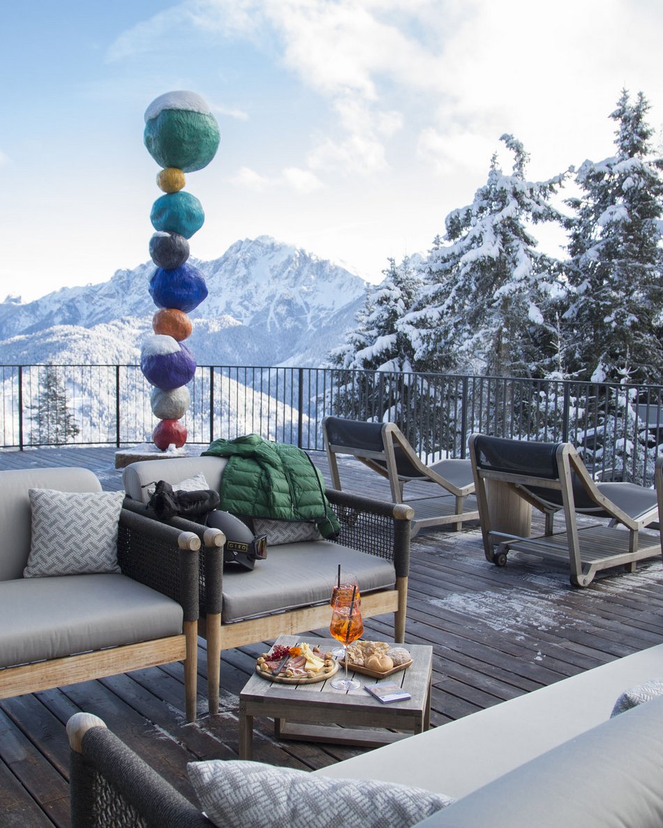 Your hotel in Val Pusteria/Pustertal: Olangerhof Terrace with sofas, snacks, and colorful sculpture in snowy mountain setting
