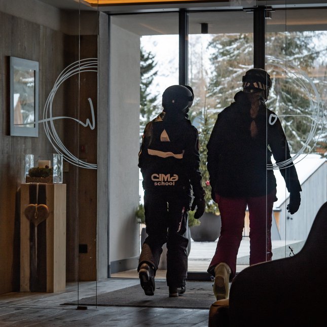 Your hotel in Val Pusteria/Pustertal: Olangerhof Two people in ski gear walking out of a building to a snowy landscape.