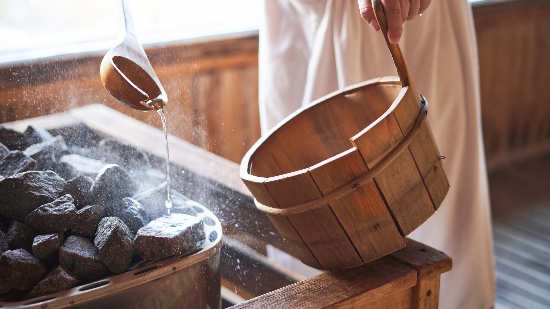 Dein Wellnesshotel am Kronplatz: Olangerhof Person gießt Wasser mit Holzlöffel auf heiße Steine in der Sauna