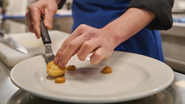 Hotels an der Piste in Südtirol mit Halbpension Koch richtet Essen in der Küche mit blauem Schürze an