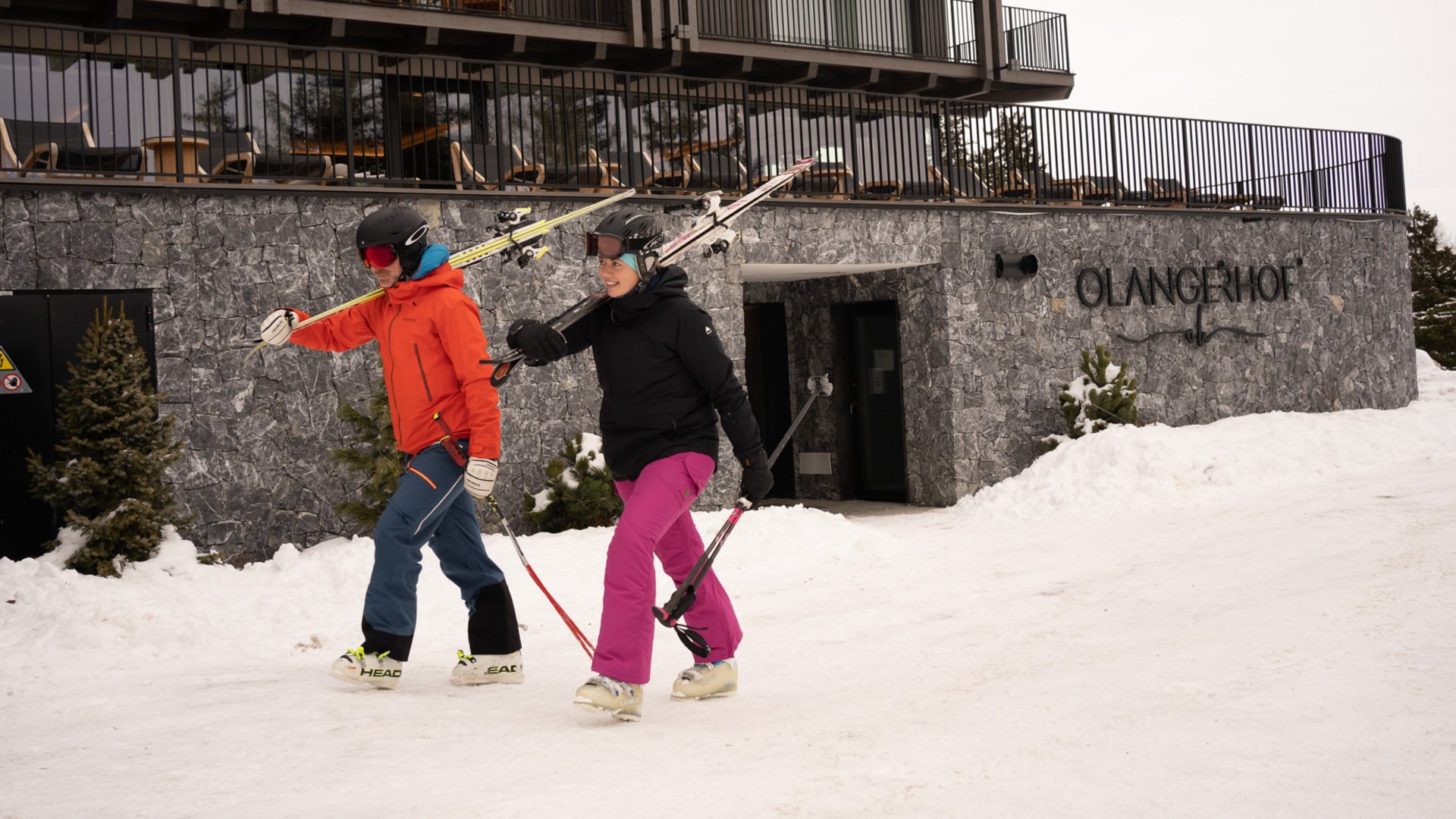 Kronplatz + Hotel an der Piste = Olangerhof Zwei Skifahrer gehen vor einem Gebäude mit dem Schriftzug Olangerhof im Schnee