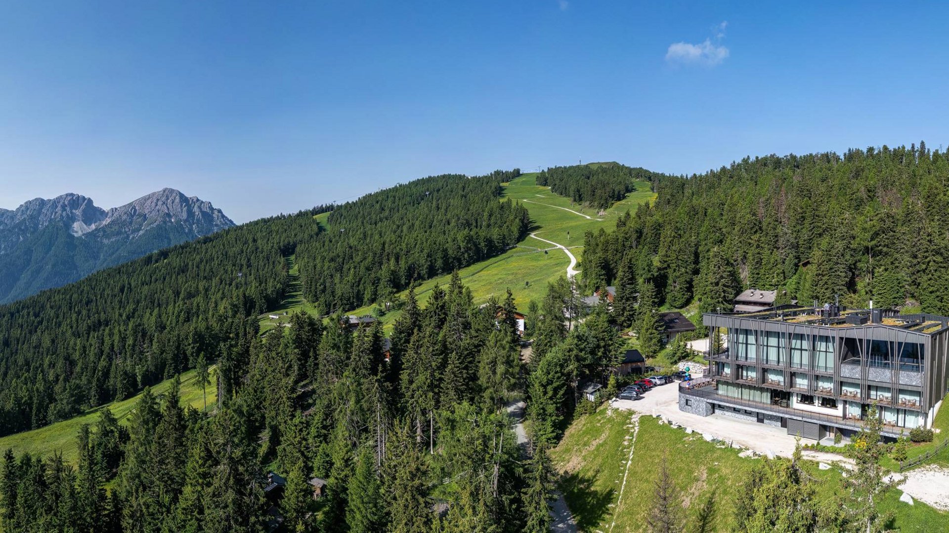 Hotels in Olang gesucht? Modernes Hotel auf grünem Hügel mit Wald und Bergen im Hintergrund