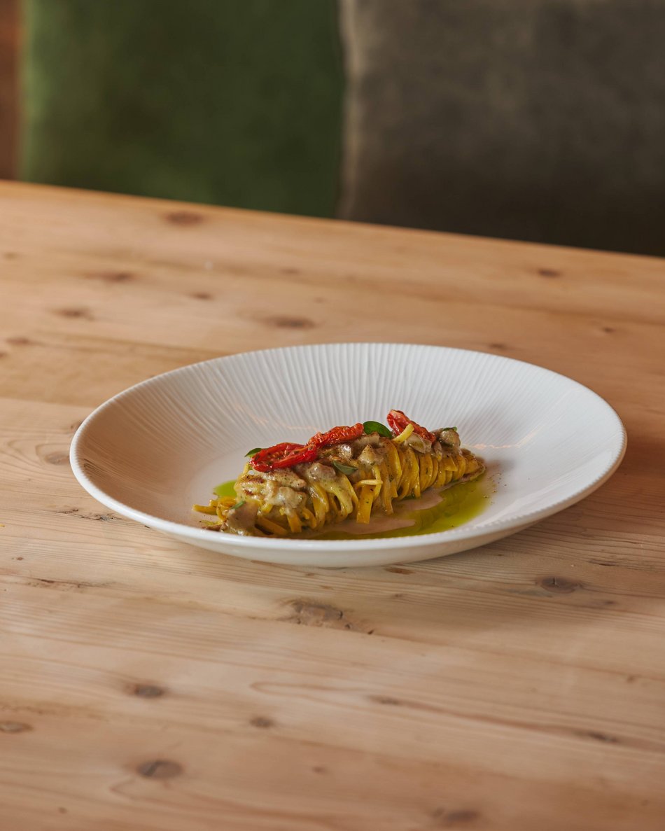 Albergo a Plan de Corones: c’è più gusto! Piatto elegante con pasta arrotolata, pomodori secchi e salsa verde su tavolo di legno