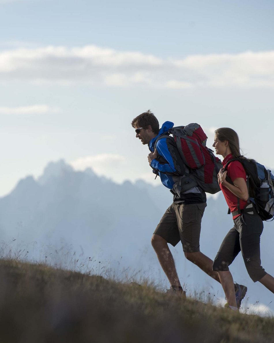 Your hotel in Val Pusteria/Pustertal: Olangerhof Two hikers with backpacks walking uphill with mountains in the background