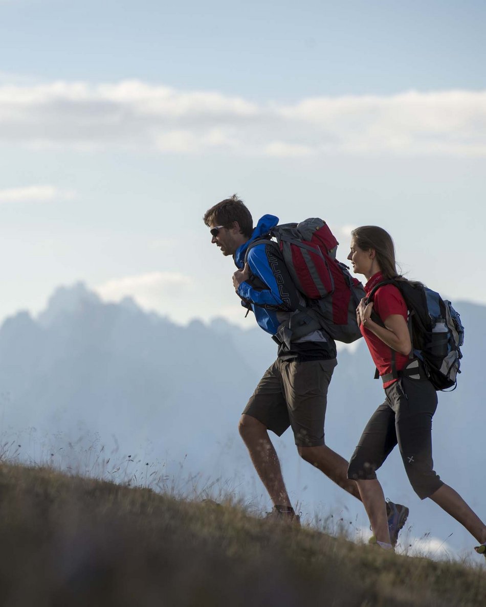 Il tuo hotel in Val Pusteria: Olangerhof Due escursionisti con zaini camminano su una collina con montagne sullo sfondo