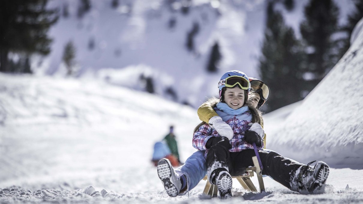 Deine Unterkunft im Pustertal: oh, wow! Zwei fröhliche Kinder rodeln mit Helm und Winterkleidung im Schnee