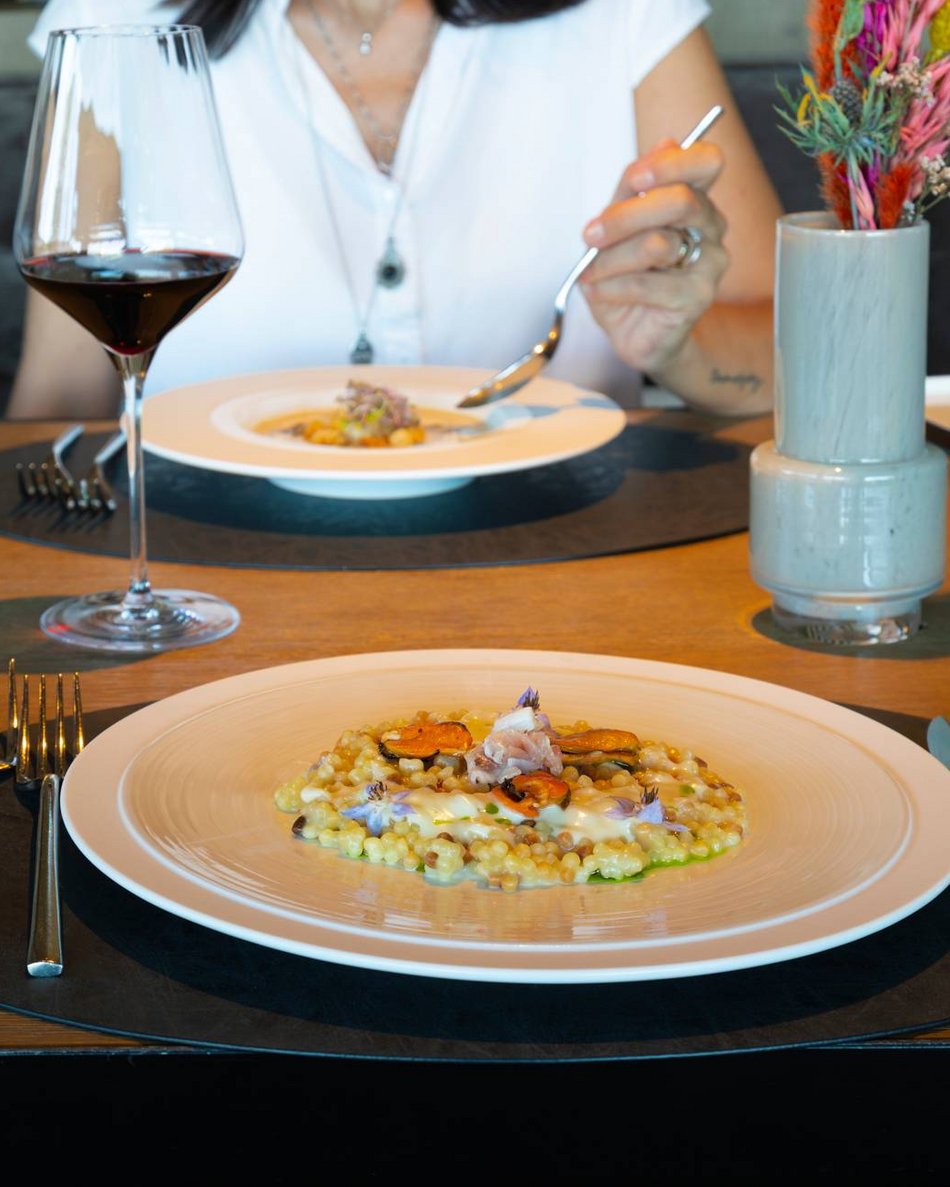 La Val Pusteria e il tuo hotel con mezza pensione: che duetto! Donna seduta a tavola con piatti gourmet e bicchiere di vino rosso