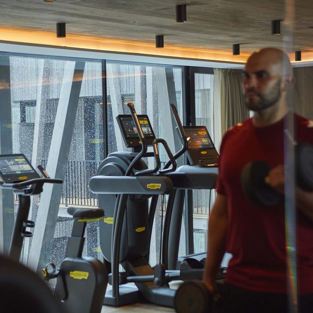 Kronplatz/Südtirol: Hotels Mann hebt Gewichte im Fitnessstudio mit Cardiogeräten im Hintergrund.