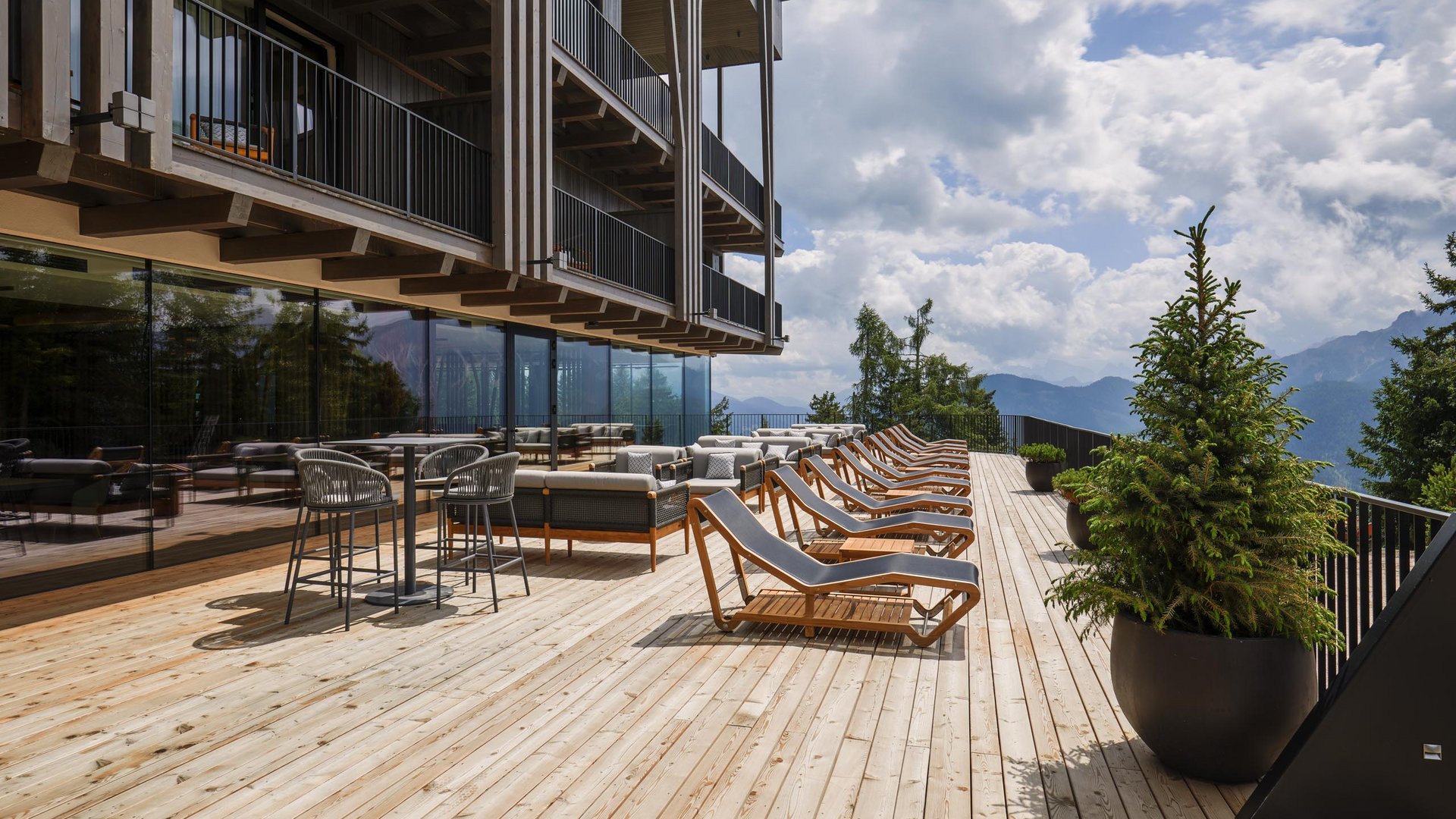 Hotels in Olang gesucht? Holzterrasse mit Liegestühlen und Tischen mit Blick auf Berge und bewölkten Himmel