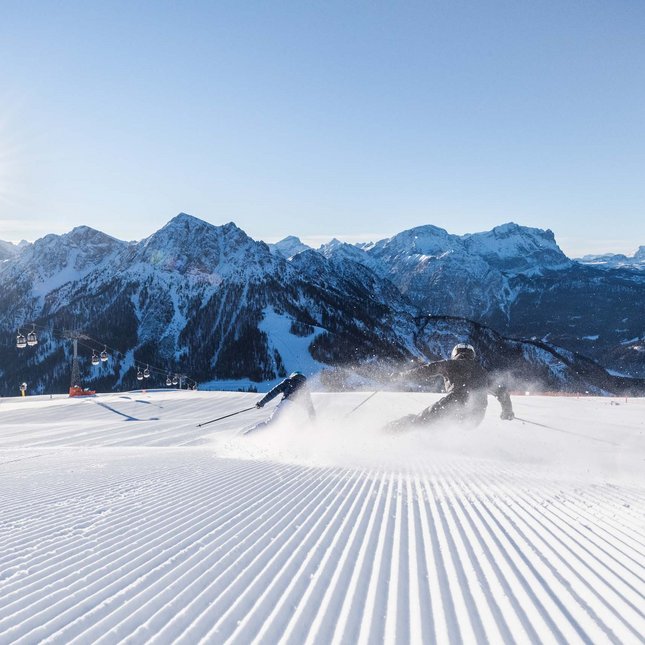 Al Plan de Corones: il tuo hotel ricco di attività Due sciatori che scendono sulla neve fresca con montagne innevate sullo sfondo