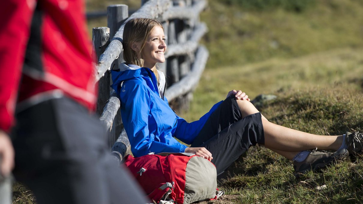 Deine Unterkunft im Pustertal: oh, wow! Frau sitzt im Gras mit blauer Jacke und rotem Rucksack neben einem Holzzaun