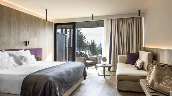 Dein Hotel in Olang: Willkommen im Olangerhof! Modernes Hotelzimmer mit großem Bett, Sofa und Balkon mit Blick auf die Bäume