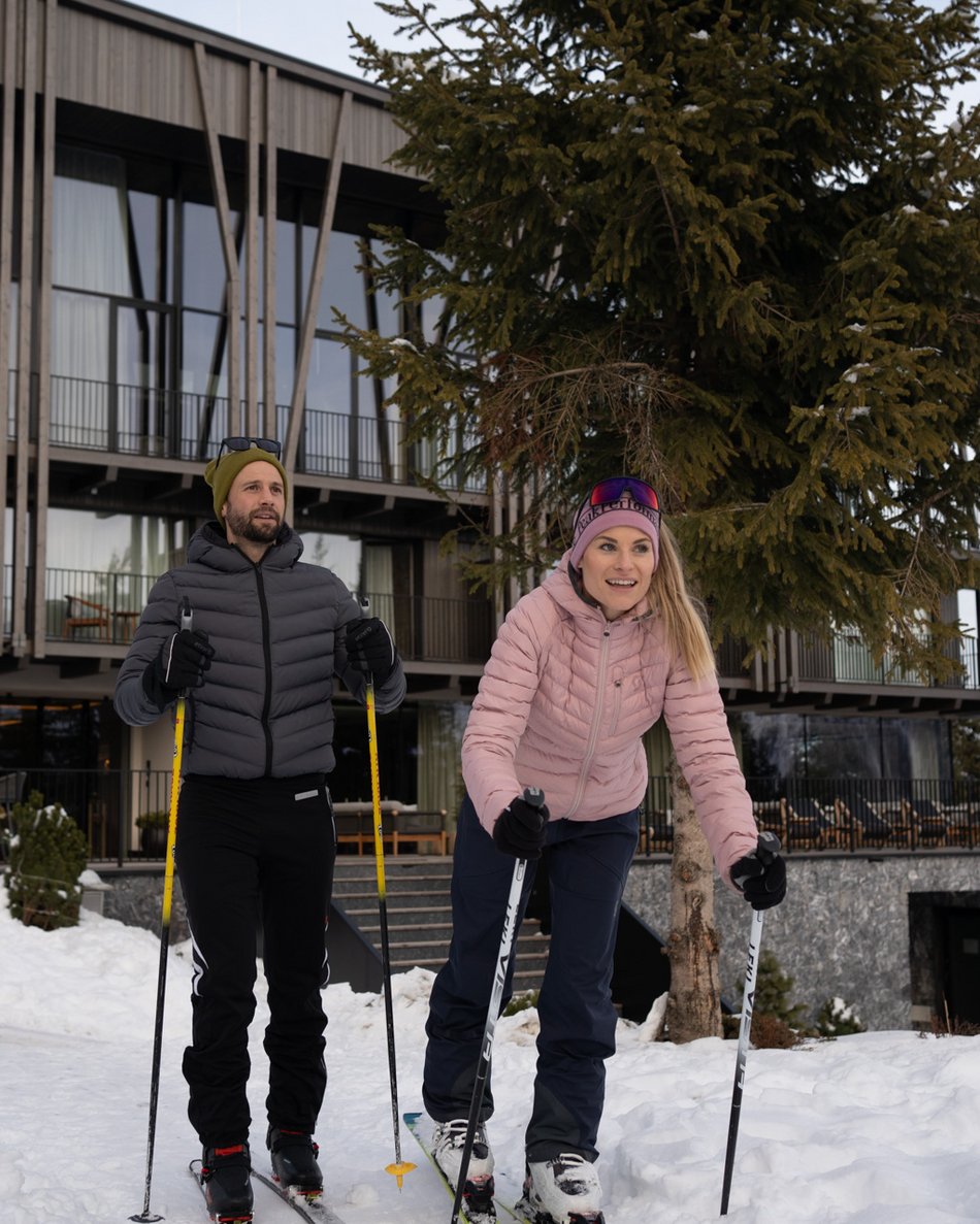 Kronplatz/Südtirol: Hotels Zwei Personen beim Langlaufen vor einem modernen Gebäude im Schnee