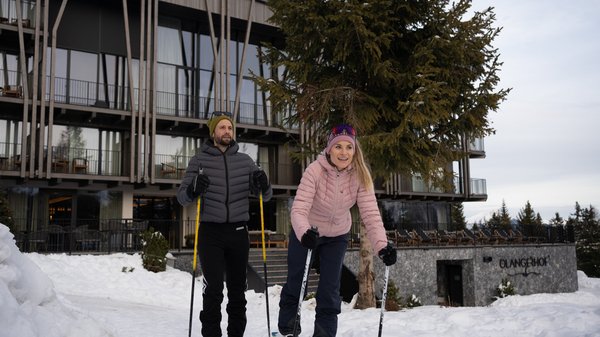 Dein Hotel in Olang: Willkommen im Olangerhof! Zwei Personen beim Langlaufen vor einem modernen Gebäude im Schnee