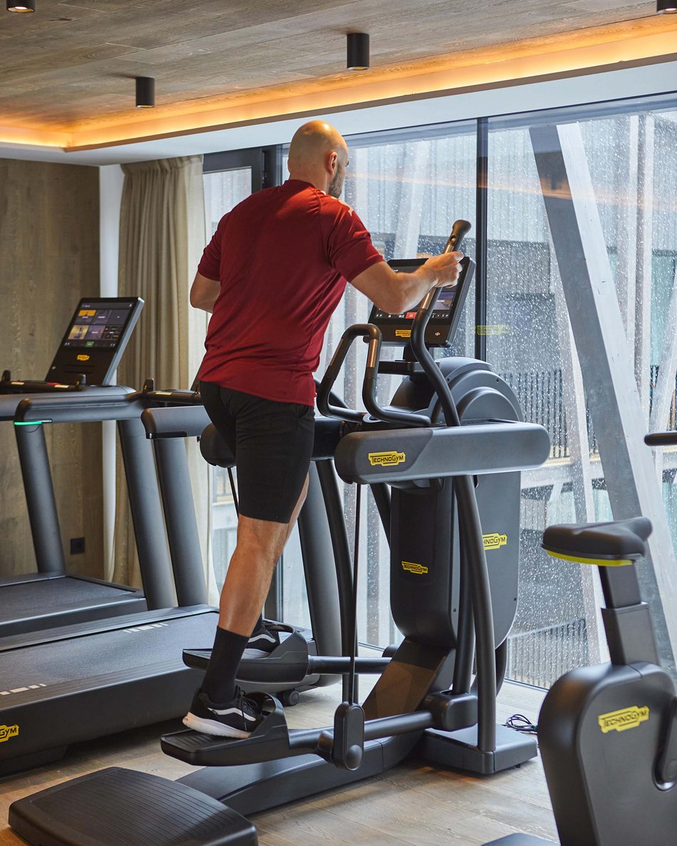 Kronplatz + Hotel an der Piste = Olangerhof Mann trainiert auf Ellipsentrainer in modernem Fitnessstudio mit Glaswand