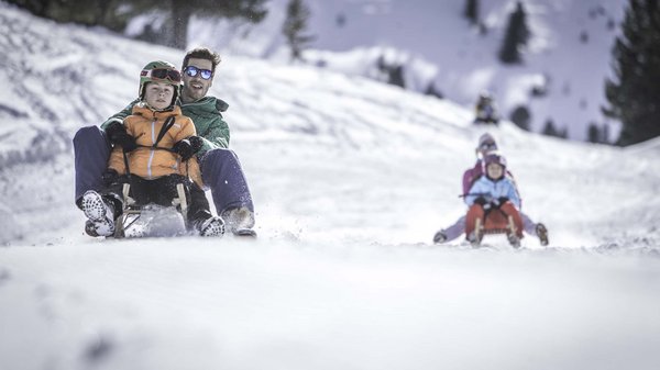 Kronplatz + Hotel an der Piste = Olangerhof Zwei Erwachsene und Kinder rodeln einen verschneiten Hügel hinunter