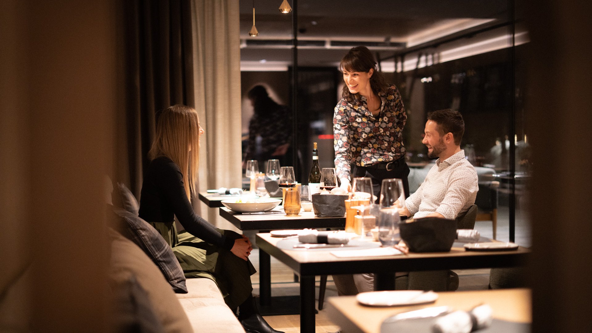 Hotels an der Piste in Südtirol mit Halbpension Kellnerin serviert zwei Gästen in einem eleganten Restaurant am Abend