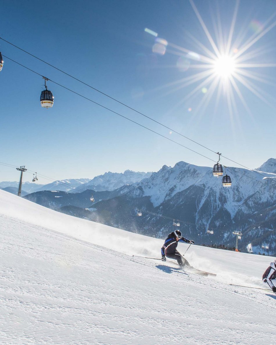 Kronplatz + Hotel an der Piste = Olangerhof Skifahrer fahren eine verschneite Piste mit Bergen und Sessellift im Hintergrund hinunter