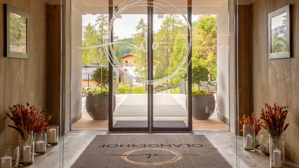Dein Hotel in Olang: Willkommen im Olangerhof! Eingang mit Holzboden, Teppich und Glastüren mit Blick ins grüne Außenbereich