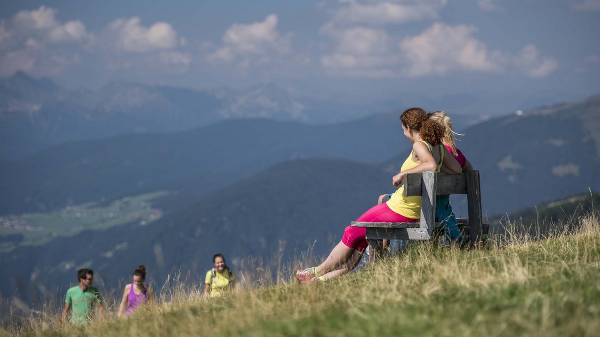 Deine Unterkunft im Pustertal: oh, wow! Zwei Frauen sitzen auf einer Bank mit Bergblick und drei Wanderer im Hintergrund