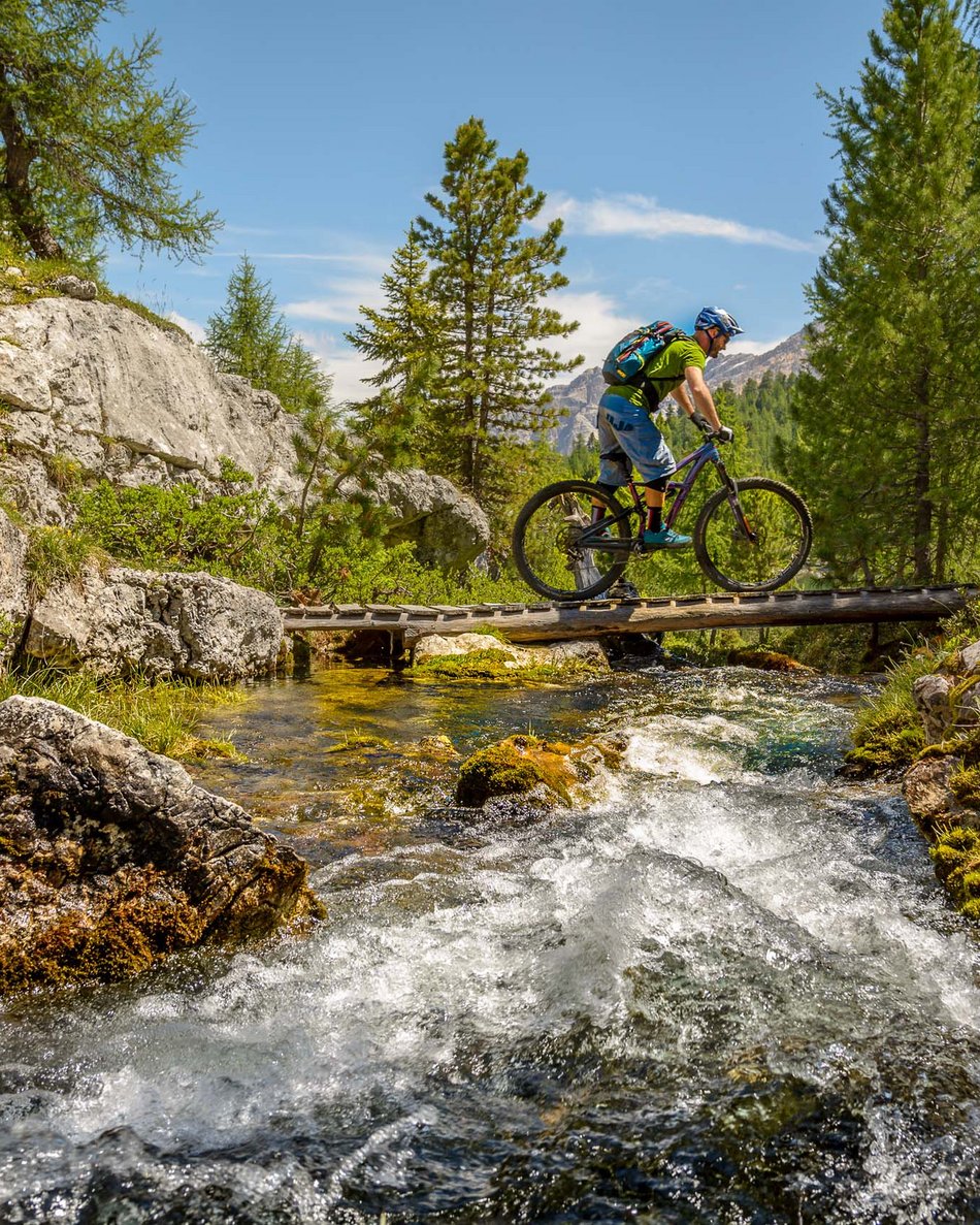 Olang in summer: sunny days at Olangerhof Cyclist wearing helmet crosses wooden bridge over mountain stream