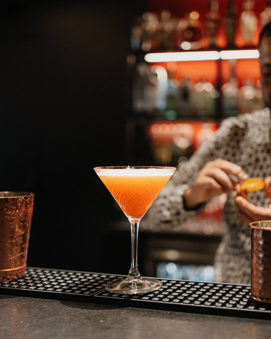 Hotel am Kronplatz mit Wine & Cocktail Bar Oranger Cocktail im Martiniglas an der Bar mit Barkeeper im Hintergrund.