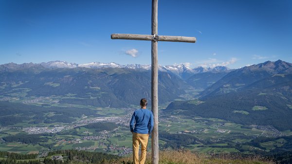 Dein Hotel in Olang: Willkommen im Olangerhof! Mann steht vor einem hölzernen Kreuz mit Panoramablick auf die Berge.