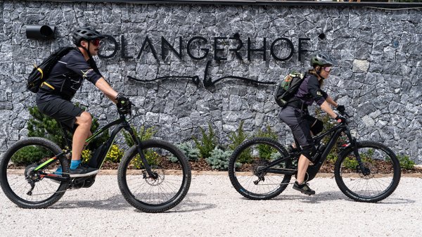 Dein Hotel in Olang: Willkommen im Olangerhof! Zwei Radfahrer mit Helm vor einer Steinmauer mit OLangerhof Schriftzug