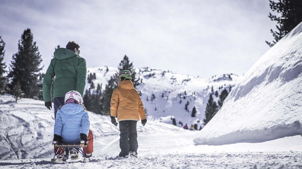 Kronplatz + Hotel an der Piste = Olangerhof Person zieht Mädchen auf Schlitten und Junge läuft im Schnee in den Bergen