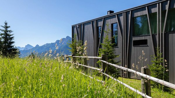 Dein Hotel in Olang: Willkommen im Olangerhof! Modernes Holzhaus mit Zaun inmitten einer grünen Wiese vor Bergen